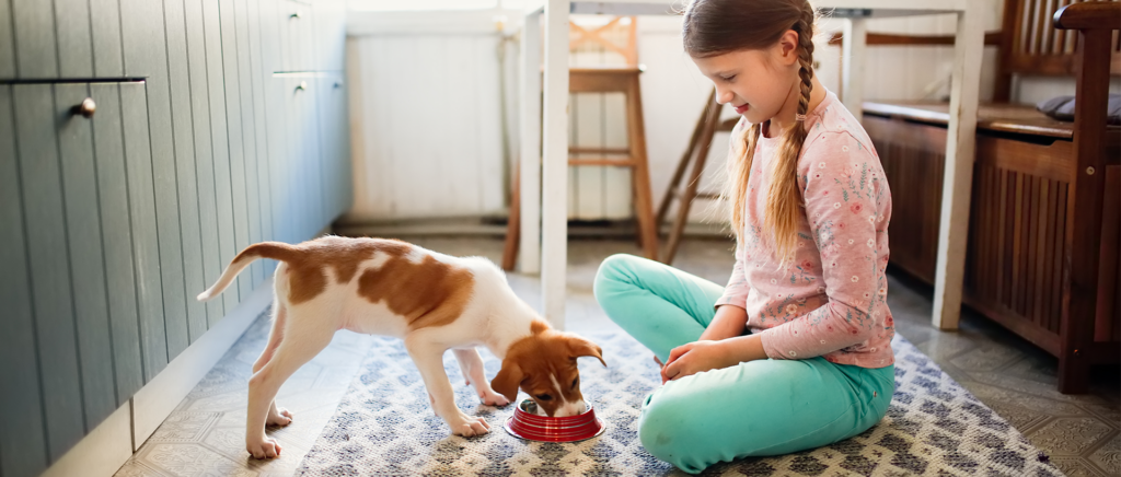 A girl sits on the floor while a puppy eats a bowl of food in front of her.